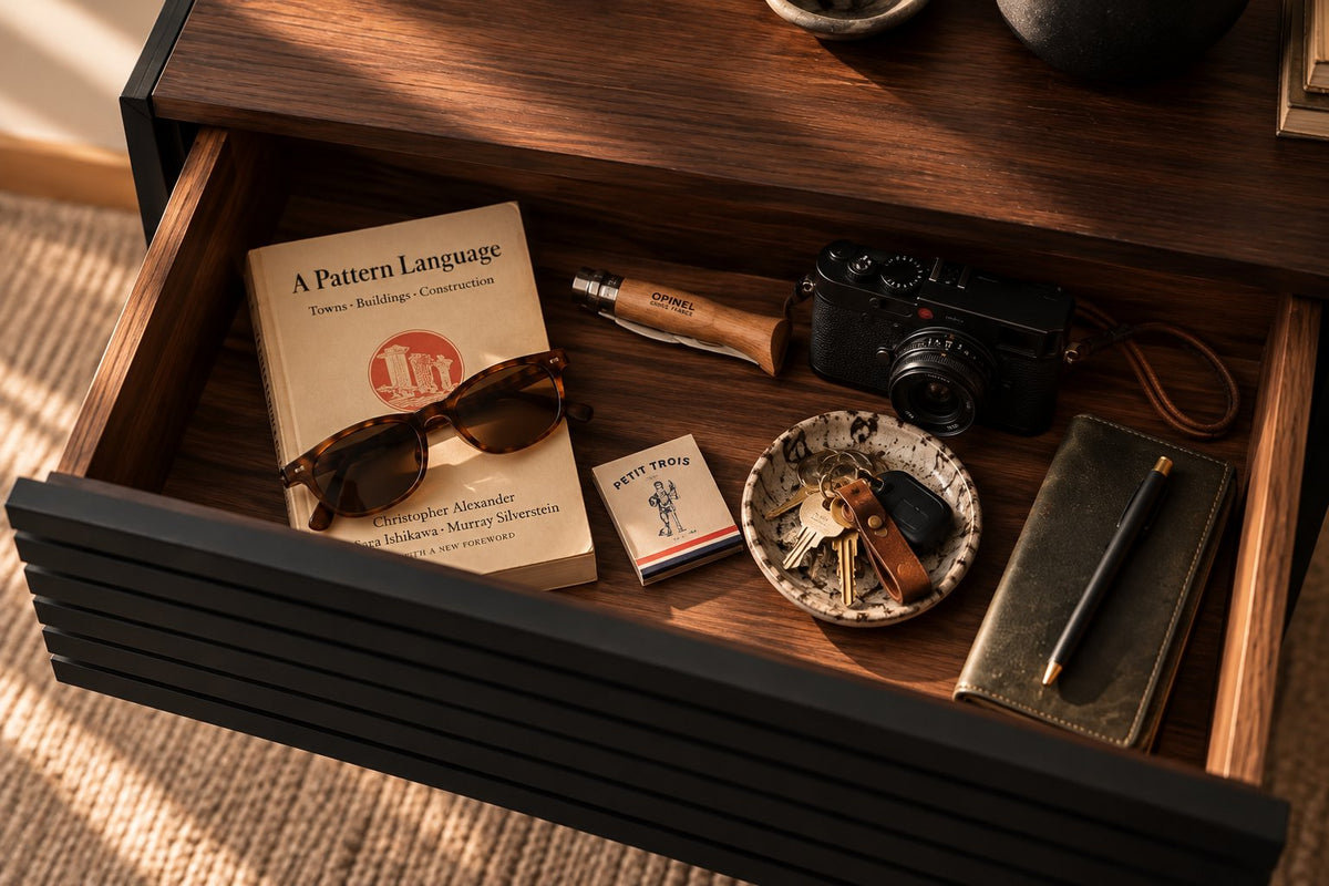 An open walnut nightstand drawer holding a book, folding knife, compact camera, sunglasses, keys and a restaurant matchbook.
