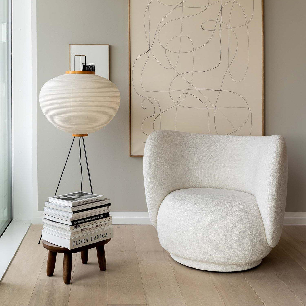 A round paper lamp on three thin legs beside a stack of books on a small wooden stool, next to a white upholstered armchair and a framed line drawing.