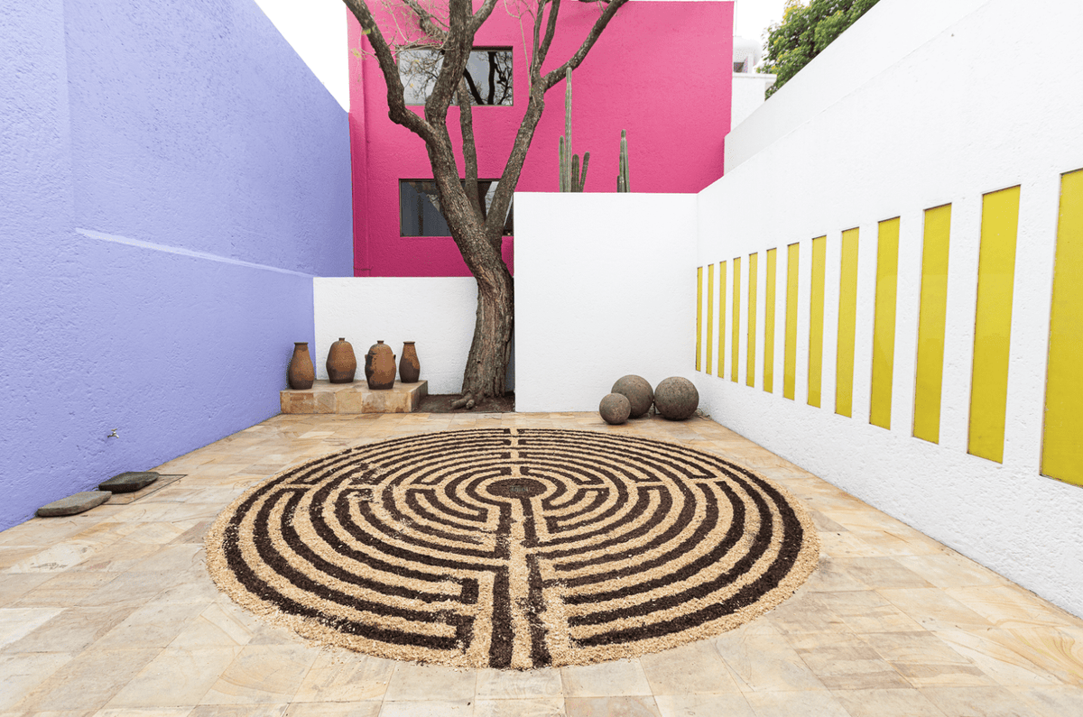 A courtyard with a lavender wall, a bright pink wall behind a tree, and a white wall set with a row of yellow vertical panels, with clay jars, stone spheres and a circular labyrinth laid out on the paving.