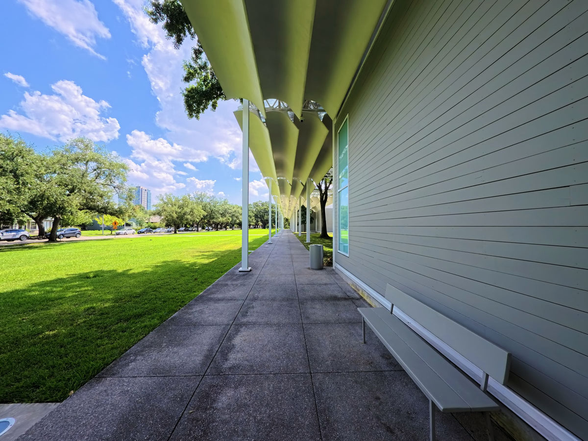 The covered walkway along the grey clapboard facade of the Menil Collection in Houston, beside a sunlit lawn.