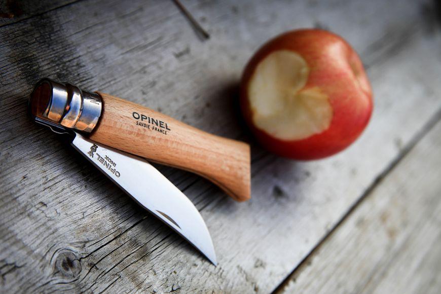 An open Opinel folding knife with a wooden handle and a steel blade marked Opinel Inox, lying on weathered grey boards beside a red apple with a bite taken out of it.