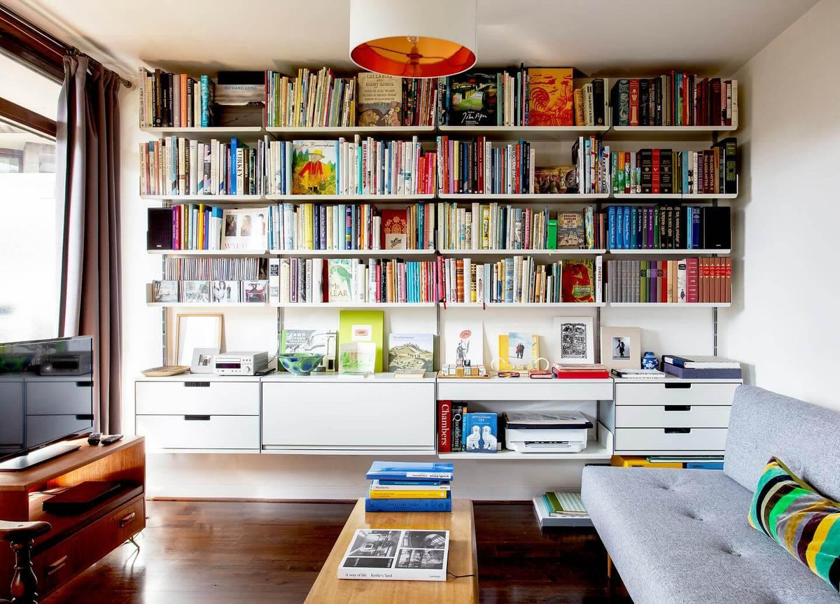 White wall-mounted shelving filled with colourful books, with drawer and cabinet units below, in a living room with a wooden coffee table and a grey sofa.
